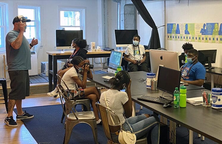Young people with cameras sitting at computer tables with a teacher giving instruction.