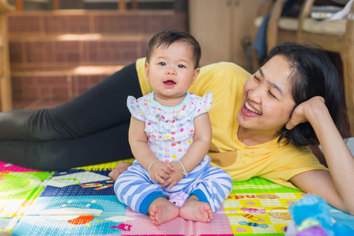 Young Asian woman lying on ground smiling at an Asian toddler that is smiling and sitting up, looking at the camera